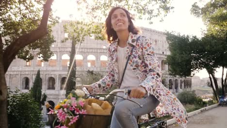 Beautiful-young-woman-in-colorful-fashion-riding-bike-in-front-of-colosseum-in-Rome-at-sunset-with-trees-happy-attractive-girl-tourist-with-straw-hat-in-colle-oppio-front-view-steadycam-dolly