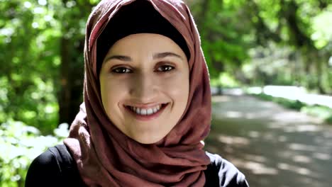 Portrait-of-a-cute-young-girl-in-a-hijab,-smiling,-looking-at-the-camera,-park-in-the-background.-50-fps