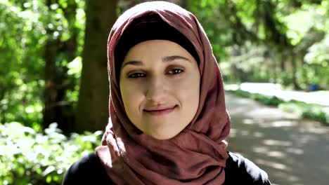 Porträt-von-einem-netten-jungen-Mädchen-in-einem-Hijab,-lächelt,-schaut-in-die-Kamera,-Park-im-Hintergrund.-50-fps