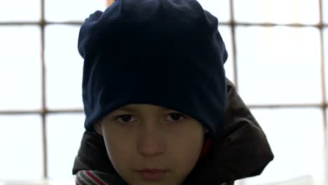 portrait-of-depressed-boy-stands-alone-near-a-broken-window,-looking-into-the-camera,-winter