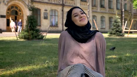 Young-beautiful-muslim-girl-dreamer-in-hijab-is-sitting-on-lawn,-builging-on-background,-religious-concept,-relax-concept