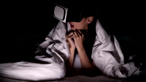 Woman-watching-VR-horror-movie-alone-in-bed.-Asian-woman-lying-in-bed-alone-wearing-VR-head-set,-very-surprise-and-shock-from-scary-movie.-High-tech-Halloween-home-alone-concept.