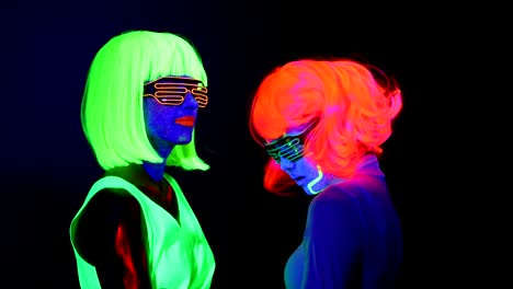 Women-with-UV-face-paint,-wig,-glowing-glasses,-glowing-clothing-dancing-together-slowly-in-front-of-camera,-Half-body-shot.-Caucasian-and-asian-woman.-.