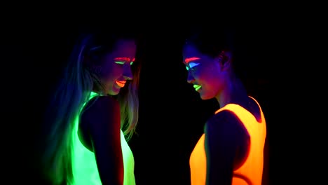 Women-with-UV-face-paint,-glowing-bracelets,-glowing-clothing-dancing-together-in-front-of-camera,-Half-body-shot.-Caucasian-and-asian-woman.-.