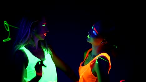 Women-with-UV-face-paint,-laser,-glowing-bracelets,-glowing-clothing-dancing-together-in-front-of-camera,-Half-body-shot.-Caucasian-and-asian-woman.-.