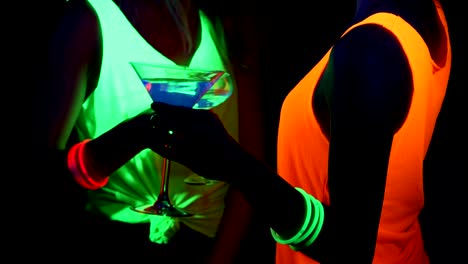 Women-with-UV-face-paint,-glowing-bracelets,-drinks,-glowing-clothing-dancing-together-in-front-of-camera,-Close-up-of-drinks.-Caucasian-and-asian-woman.-.