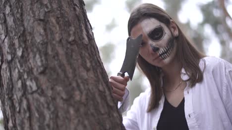 Halloween.-Mädchen-mit-einem-Messer-und-Halloween-Make-up-versteckt-sich-hinter-einem-Baum