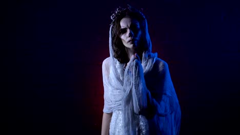 Ghost-of-lonely-dead-bride-in-white-wedding-dress-and-veil-standing-with-sad-face-moving-her-hand.-Scary-girl-halloween-apperance-make-up-against-horror-background.-Mysterious-woman