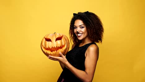 Funny-african-lady-in-halloween-costume-showing-and-playing-with-pumpkin-isolated-over-yellow-background