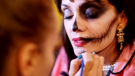 Halloween-party,-make-up-artist-draws-a-terrible-makeup-on-the-face-of-a-brunette-woman-for-a-Halloween-party.-in-the-background,-the-scenery-in-the-style-of-Halloween-is-seen