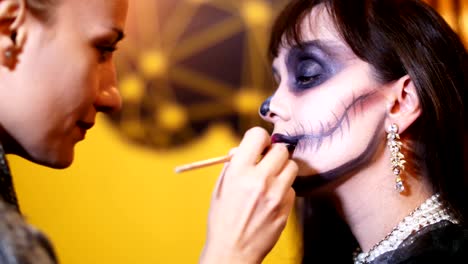 Fiesta-de-Halloween,-Maquillaje-artista-dibuja-un-maquillaje-terrible-en-la-cara-de-una-mujer-morena-para-una-fiesta-de-Halloween.-en-el-fondo,-se-ve-el-paisaje-en-el-estilo-de-Halloween