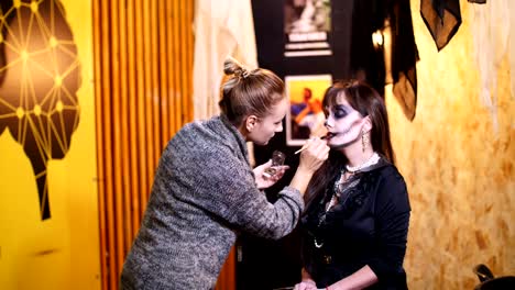 Fiesta-de-Halloween,-Maquillaje-artista-dibuja-un-maquillaje-terrible-en-la-cara-de-una-mujer-morena-para-una-fiesta-de-Halloween.-en-el-fondo,-se-ve-el-paisaje-en-el-estilo-de-Halloween