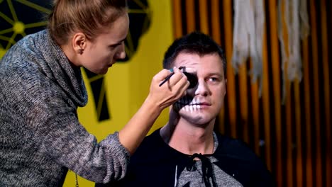 Halloween-party,-make-up-artist-draws-a-terrible-makeup-on-the-face-of-a-man-for-a-Halloween-party.-in-the-background,-the-scenery-in-the-style-of-Halloween-is-seen