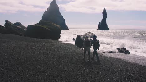 Young-couple-holding-hands-walking-down-black-sand-beach-in-Iceland,-slow-motion