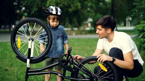 Neugieriges-Kind-tragen-Helm-dreht-Rad-und-Pedale,-während-sein-Vater-auf-Rasen-im-Park-am-Sommertag-im-Gespräch-mit-ihm-ist.-Familie,-Freizeit-und-aktiven-Lifestyle-Konzept.