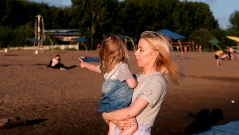 Kleines-Mädchen-in-der-Mutter-Armen-zeigt-etwas-mit-der-Hand-auf-die-Seite-am-Strand-am-Fluss.