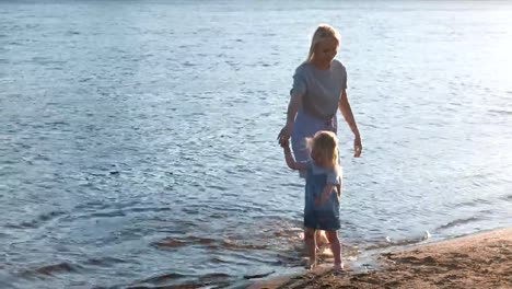 Mom-and-girl-walking-on-the-beach-of-the-river-at-sunset-and-swim-in-city.