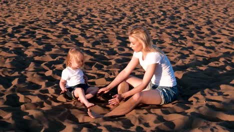 Schöne-blonde-Mutter-und-Tochter-spielen-mit-Sand-sitzen-zusammen-am-Strand.