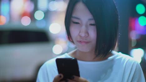 Portrait-of-the-Attractive-Japanese-Girl-Wearing-Casual-Clothes-Uses-Smartphone.-In-the-Background-Big-City-Advertising-Billboards-Lights-Glow-in-the-Night.
