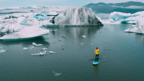 Toma-aérea-del-hombre-remando-de-pie-Junta-de-paleta-en-la-laguna-de-glaciar-con-icebergs-gigantes-en-Islandia