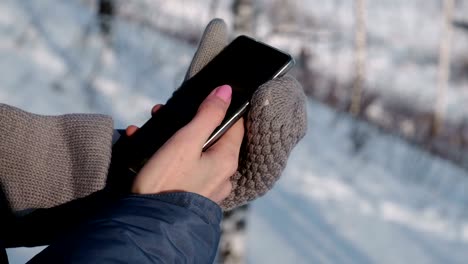 Close-up-woman's-hands-in-mittens-writing-messages-on-cellphone-in-winter-day-in-park.