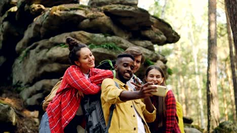 Happy-African-American-man-tourist-is-taking-selfie-with-friends-in-forest-near-huge-rocks-using-smartphone,-people-are-making-funny-faces-and-showing-hand-gestures.