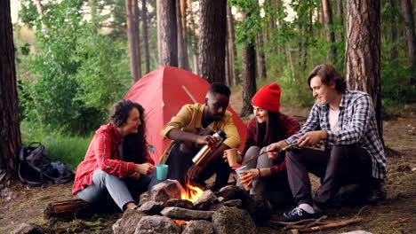 Guapo-hombre-afroamericano-es-verter-bebida-caliente-de-termo-en-vasos-y-copas-sentados-alrededor-del-fuego-en-el-campamento-con-grupo-de-amigos-de-multiétnico.