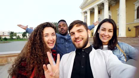 Point-of-view-shot-of-multiracial-group-of-tourists-taking-selfie-in-city-center-holding-camera-and-posing-together-with-hand-gestures-expressing-positive-emotions.