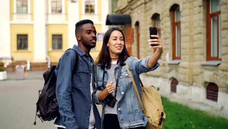 Schöne-gemischtrassige-Paar-ist-im-Gespräch-mit-Freunden-online-video-Anruf-mit-Smartphone-stehen-auf-der-Straße-mit-Rucksäcken.-Reisen-und-Technik-Konzept.