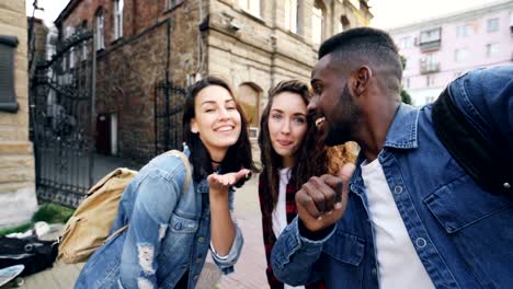 Point-of-view-shot-of-attractive-young-men-and-women-travelers-taking-selfie-in-the-street-posing-and-laughing-holding-camera.-Modern-technology-and-travelling-concept.