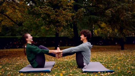 Glückliche-junge-Frauen-tun-Paar-Yoga-mit-Spaß-und-Lachen-auf-Matten-auf-dem-Rasen-im-Park-sitzen.-Schönen-Herbst-Natur-Rasen-und-Bäume-sind-im-Hintergrund.