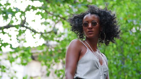 Trendy-young-black-woman-in-camisole-looking-up-at-trees-turns-to-the-camera,-low-angle,-waist-up