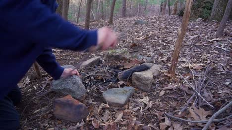 Man-builds-campfire-to-cook-pot-over-fire.-Essential-Bushcraft-/-Survival-skill.-Primitive-debris-hut-shelter-at-the-camp-site.-Camping-overnight-in-the-blue-ridge-mountains-of-North-Carolina