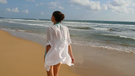 Schöne-Frau-im-Badeanzug-und-Hemd-zu-Fuß-am-Strand-und-Hände-erhoben.-Junge-Mädchen-gehen-direkt-am-Ozean-und-Sommer-genießen.-Wellen-des-Meeres-im-Hintergrund.-Urlaub-Konzept-zurück-Rückansicht