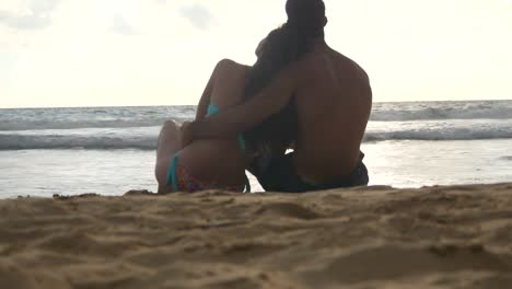 Young-romantic-couple-is-enjoying-beautiful-view-sitting-on-the-beach-and-hugging.-A-woman-and-a-man-sits-together-in-the-sand-on-the-seashore,-admiring-the-ocean-and-landscapes.Close-Up