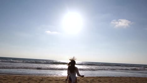 Young-woman--running-on-beach-to-the-ocean-at-sunset-and-raised-hands.-Beautiful-young-girl-going-on-sandy-shore-to-the-sea-and-enjoying-freedom-during-vacation.-Relax-on-summer-holiday.-Rear-back-view