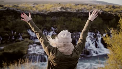 Libertad:-mujer-joven-de-pie-cerca-de-cascadas-de-Islandia-y-levantando-las-manos,-lleva-sombrero-y-esta-feliz