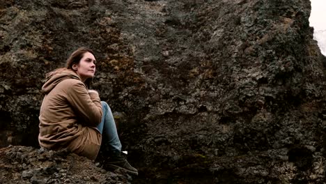 Young-beautiful-woman-sitting-on-the-rock-in-mountains-and-thinking-alone.-Tourist-resting-after-hiking-in-Iceland