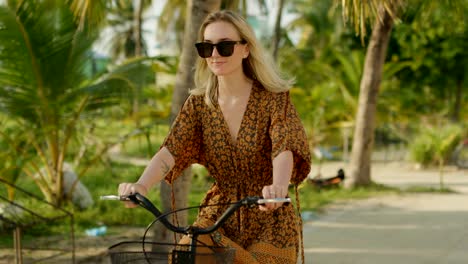 Beautiful-Woman-Wearing-Dress-Rides-Bicycle-on-the-Road-Through-Authentic-Small-Town.-South-Asia-Nature,-Palm-Trees-Grow-by-the-Road.-Close-up-Slow-Motion.