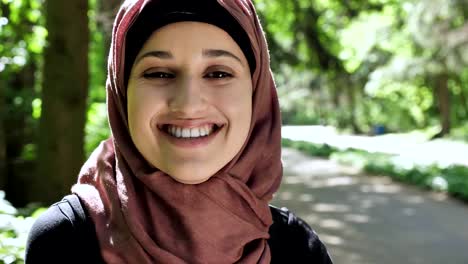 Portrait-of-a-cute-young-girl-in-a-hijab,-smiling,-looking-at-the-camera,-park-in-the-background.-50-fps