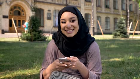 Joven-hermosa-chica-musulmana-en-hijab-es-viendo-en-smartphone-en-el-día-en-verano,-mirando-a-cámara,-Sonriente,-basándose-en-antecedentes,-concepto-de-epoca,-concepto-de-comunicación