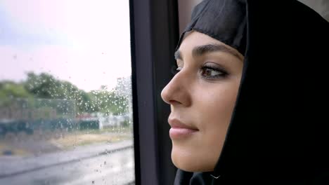 Young-muslim-woman-in-hijab-is-watching-in-rainy-window-in-bus,-transport-concept,-urban-concept,-weather-concept,-dreaming-concept,-side-view