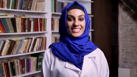 Young-happy-muslim-woman-in-hijab-standing-in-library-and-looking-at-camera-and-smiling