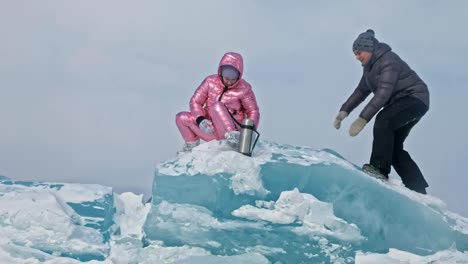 Young-couple-has-fun-during-winter-walk-against-background-of-ice-of-frozen-lake.-Lovers-are-sitting-on-large-blue-ice-floe,-kissing-and-hugging,-drinking-tea-from-thermos-bottle.-Love-story.