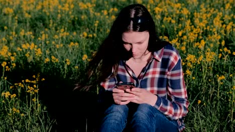 Frau-Handy-sitzen-im-Park-auf-dem-Rasen-unter-den-gelben-Blüten-betrachten.