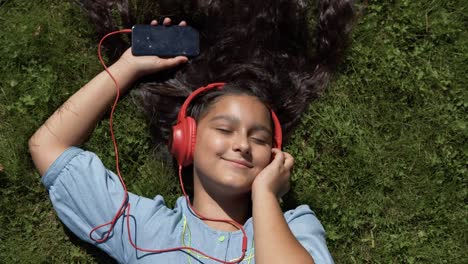 Junges-Mädchen-mit-langen-schwarzen-Haaren,-anhören-von-Musik-über-Kopfhörer-bei-sonnigem-Wetter-auf-dem-Rücken-im-Park-liegt.-4K