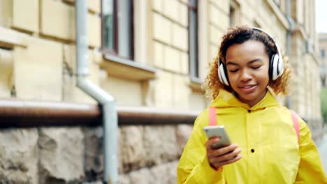 Feliz-chica-afroamericana-está-escuchando-música-con-auriculares-inalámbricos-y-utilizando-smartphone-caminando-al-aire-libre-disfrutando-de-la-canción-y-a-pie.-Concepto-de-gadgets-y-Milenio.