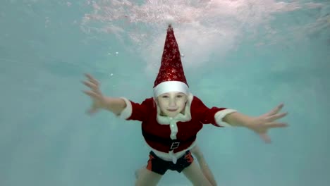 Glücklicher-kleine-Junge-in-einem-roten-Anzug-Weihnachtsmann-schwimmt-unter-Wasser-im-Pool-in-den-Wasserstrahl,-lächelnd-und-Blick-in-die-Kamera.