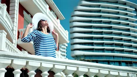 Elegant-woman-in-hat-and-sunglasses-raising-hands-enjoying-sun-standing-on-balcony