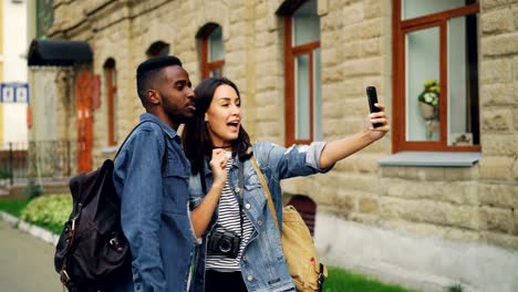 Excited-tourists-man-and-woman-are-making-online-video-call-with-smartphone-holding-device-and-talking-showing-thumbs-up-expressing-positive-emotions.